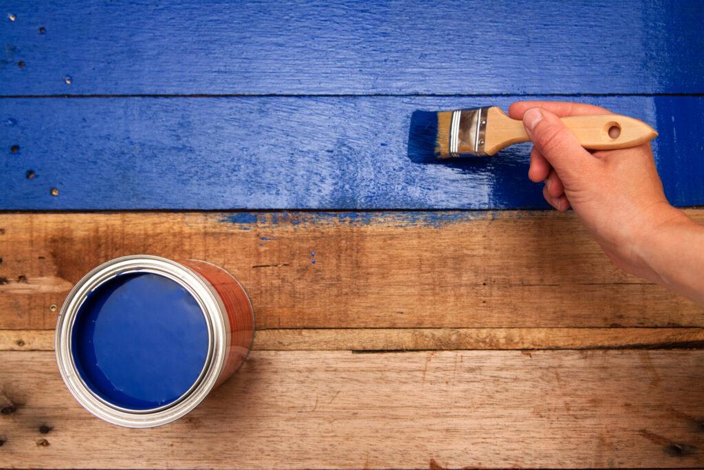 Hand streicht mit Pinsel und blauer Farbe einen hölzernen Hintergrund