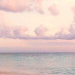 Frau im weißen Hochzeitskleid, die bei Sonnenuntergang am Strand mit pastellfarbenem Himmel und Ozean spazieren geht