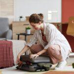 Junge Frau kniet auf dem Boden beim Kofferpacken, vorbereitet auf eine Wellnessreise mit Duschtuch mit Namen